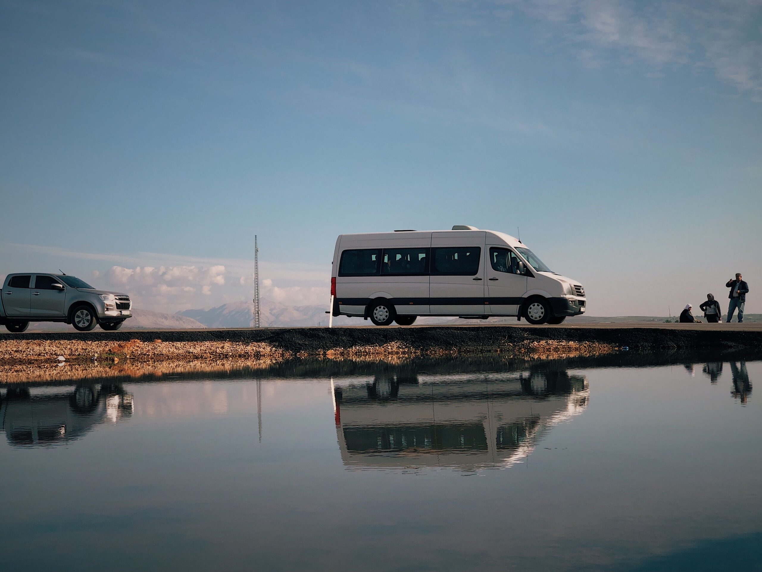 A van and trucks parked by a tranquil lakeshore in Adıyaman, Turkey, beautifully reflected in still water.